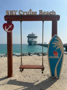 Photo of a wooden swing on a beach with reads 'SBY Cruise Beach' and cruise ship in the background at sea 